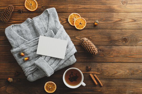 Top view cozy home wooden desk with knitted sweater, tea cup, blank paper card, dry oranges, acorns, pine cones. Flat lay, top view. Nordic, hygge, comfort concept.