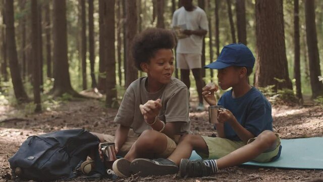 Slowmo shot of two African American siblings sitting on forest ground at campsite snaking with tea and sandwiches while their dad observing map in background
