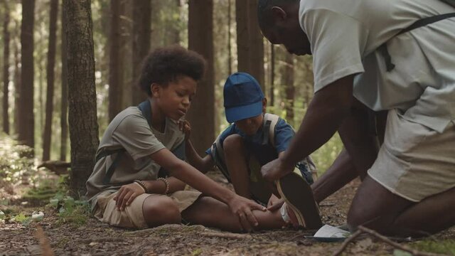 Slowmo shot of African American boy resting on forest ground after having injured his leg while hiking together with dad and little brother. Man examining his ankle