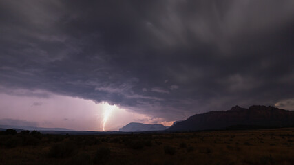 Fototapeta na wymiar A bolt of lightning drops down from the edge of rolling purple clouds in a landscape with an open grassy field dotted with sagebrush and the mountains of Southern Utah in the distance. 