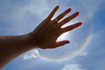 Woman's hand with ring and painted nails covering the sun in a solar halo effect, on a celestial sky.