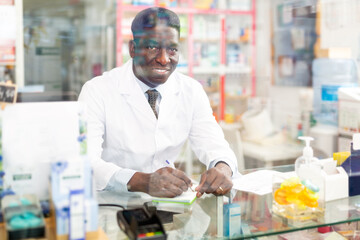 Fototapeta na wymiar Smiling african american male pharmacist working in a pharmacy makes important notes while sitting behind the counter