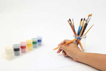 Hand of person holding pencil before drawing on white sheet with brushes and tempera around....