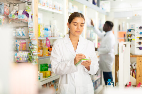 Portrait of a focused young female pharmacist standing in the sales hall of a pharmacy, making important work notes in a ..notebook