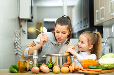 Positive mother and little daughter tasting soup together at home