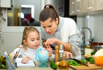 Positive mother cooking and little daughter doing school homework at kitchen