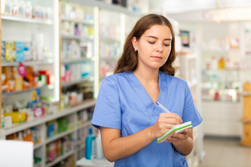 Female pharmacist standing with notebook in hands in salesroom of drugstore
