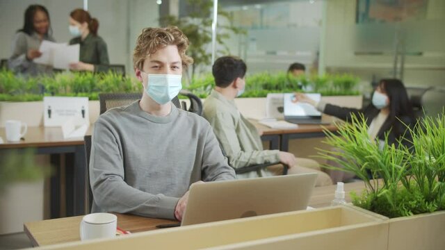 A young coworker is sitting in a facial protective mask and looking to the camera