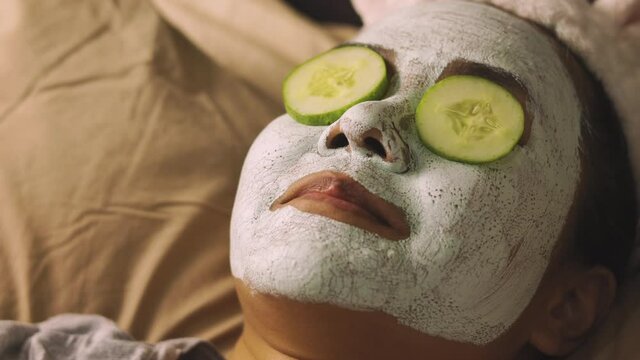 Face focusing of medium aged asian woman who is masking face with mud and sliced cucumber on her eyes on bed with relaxing warm light and listening music for happy and enjoy relaxing moment.
