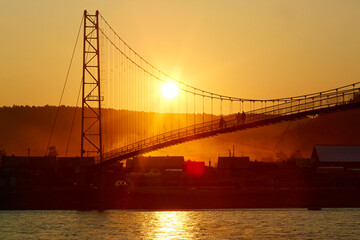 Fototapeta na wymiar People walk on a suspension bridge over the river at sunset