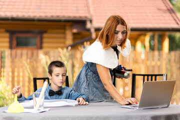 Mom and boy schoolboy are engaged in lessons through a laptop at home in the garden. Online classes for children. Schoolboy listens to a lecture and solves problems