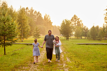 Naklejka na ściany i meble Content loving father walking with preteen daughters along green meadow in village in sunset