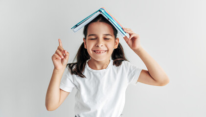 Studio portrait of a pretty little girl smiling with closed eyes holding book over head like roof pointing up with index finger, isolated over grey background.