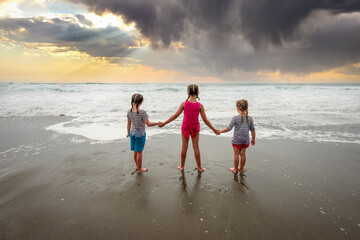 Three girls holding hands at the beach looking at the ocean and beautiful sunset - 452646041