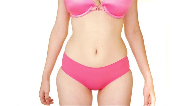 With an uncomfortable expression, a woman wearing a pink bra and underwear, trying to hide her tummy with her two hands. A sexy woman standing enjoy touching her slim tummy with her hands.