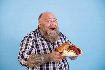 Happy emotional plump man with open mouth holds plate of different fat food posing on light blue...