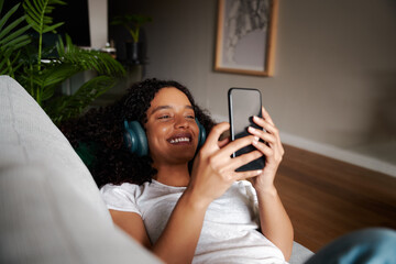 Mixed race female student lying on couch texting on cellular device listening to music