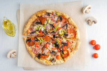 Top view of one whole sliced tasty italian pizza made of mushrooms, tomatoes, ham, pepper and cheese served on craft brown paper with olive oil on white wooden background. Horizontal image