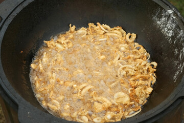Fresh onion slices are fryng in boiling oil in black kazan, closeup view. Many ruddy tasty pieces of onion are frying deep-fried in the cauldron in wood-burning stove on nature, closeup top view.