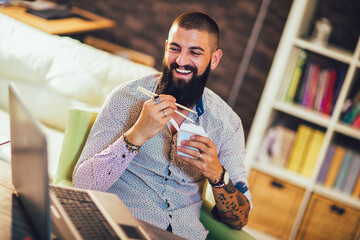 Young man working on his laptop computer at home and eating Chinese food