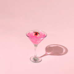 Eyeball figurine with eyelash in martini cocktail glass on pastel pink background. Halloween minimal creative concept. Pink eye retro fashion aesthetic idea.