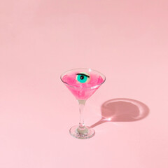 Eyeball figurine with eyelash in martini cocktail glass on pastel pink background. Halloween minimal creative concept. Blue eye retro fashion aesthetic or cosmetic idea.