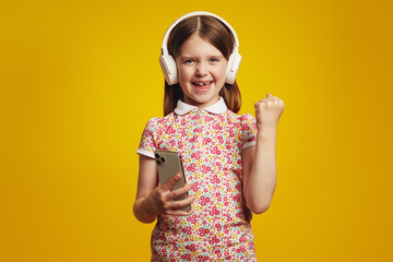 Happy surprised young girl kid in summer clothes and headphones isolated over yellow wall background using mobile phone make winner, yes gesture