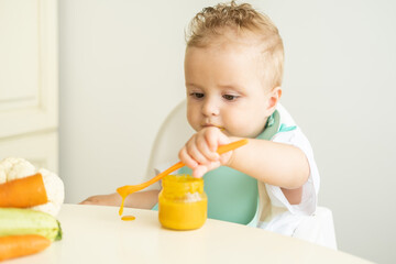 funny baby boy in bib eating vegetable puree with spoon sitting in child chair. Child learn to eat by himself