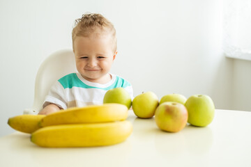 Fototapeta premium cute baby boy sitting at the table in child chair eating apple on white kitchen.