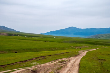 Fototapeta na wymiar Gravel road on green mountain plateau with mountains on background. Assy plateau, Almaty region, Kazakhstan.Travel, tourism in Kazakhstan concept. Nature landscape.