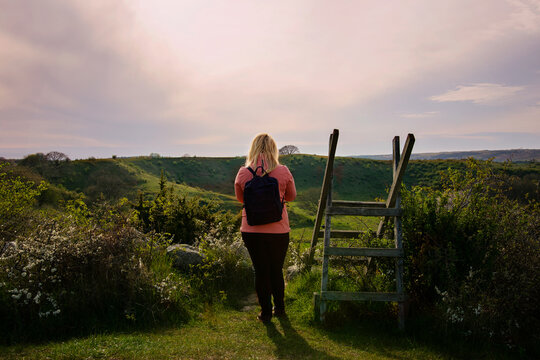 Woman walking on hiking trail during sunset in countryside landscape in Sk&aring;ne, Sweden. Woman wearing outdoor clothing with a pink wind jacket and a backpack. Photo taken at Br&ouml;sarps Backar, &Ouml;sterlen