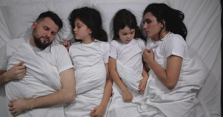 Top view of Caucasian family with two children sleeping in bed with white sheets. High angle shooting. A family of four is sleeping. Mom, dad and daughters good night, restful sleep 4k