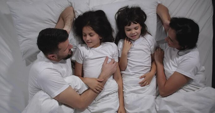 Top view of Caucasian family with two children laughing and talking in bed with white sheets. High angle shooting. A family of four is sleeping. Mom, dad and daughters good night, restful sleep 4k