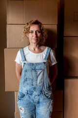 Portrait of mature adult blonde woman looking at camera wearing jean overalls in front of pile of...