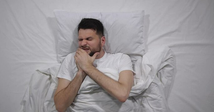  Top view  of caucasian bearded man lying in bed with white linens in light room suffering from headache. Attractive guy suffers from pain migraine holding head with hands   4K 