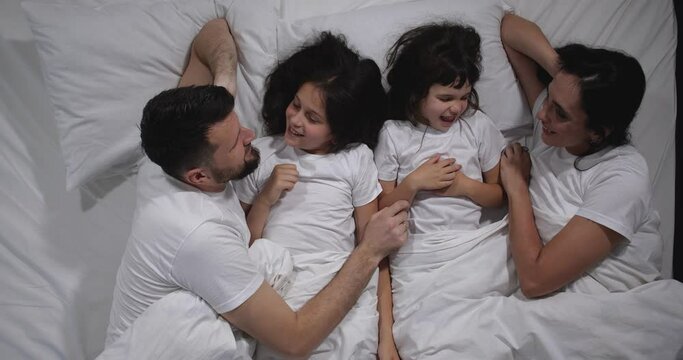 Top view of Caucasian family with two children laughing and talking in bed with white sheets. High angle shooting. A family of four is sleeping. Mom, dad and daughters good night, restful sleep 4k