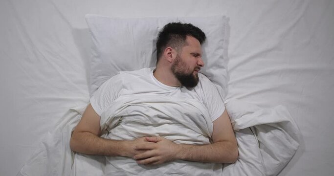  Top view  of caucasian bearded man lying in bed with white linens in light room suffering from toothache. Attractive guy suffers from dental  pain  holding face with hands   4K 