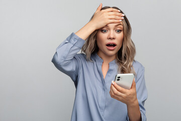 Naklejka premium Shocked young woman read stressful or surprising message in smartphone holding hand on forehead worried with mouth open in shock. Isolated studio shot of amazed girl get email or text in mobile phone