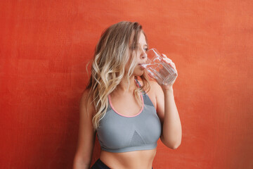 A young caucasian charming slim blonde woman with wavy hair in sports clothes drinks water from a large transparent glass after training standing against an orange wall. Morning routine, wellness