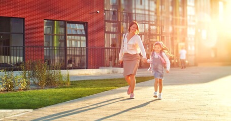 Caucasian happy mother and cute daughter walking outdoors on sunny day from school after lessons. Pretty joyful woman with little schoolgirl jumping and smiling on street. Education concept