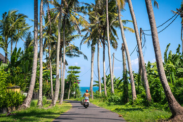 Surfer girl in swim suit rides motorbike on road to the ocean with palm trees i