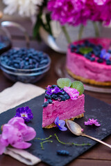 Fototapeta premium A piece of berry cheesecake lying next to a mug of tea, violet flowers and on a background of peonies and a whole cake
