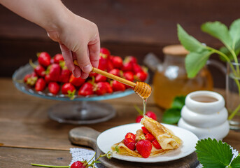 Thin pancakes stacked with strawberries between layers lying on a plate. Hand holding honey spoon and honey flowing on a stack of pancakes