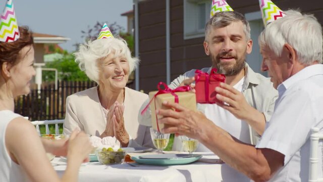Happy family members in party hats saying congratulations and giving birthday presents to grandfather and then applauding while having outdoor celebration dinner on summer day