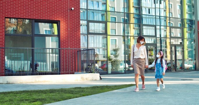 Caucasian Beautiful Parent And Cute Daughter Pupil In Masks Walking Outdoors On Sunny Day. Woman Walks With Little Junior Student From School In Quarantine And Talking. Back To School Concept