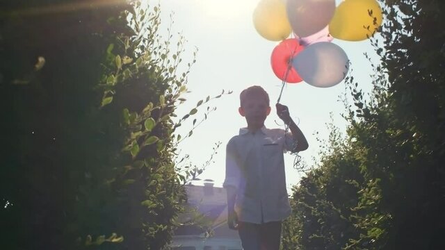 Little caucasian boy holding bunch of colorful balloons and walking through green bushes towards the camera on sunny summer day