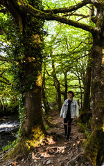 Attractive young female wearing a hat and denim jacket while walking between trees in a rain forest in Cornwall, United Kingdom
