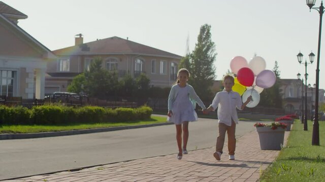 Happy little kids holding hands and walking together along the street on summer day. Cheerful girl jumping and boy holding colorful air balloons