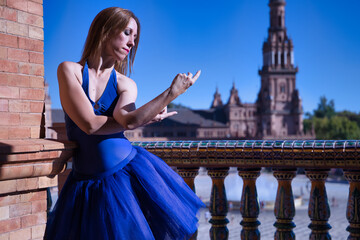 Hispanic adult female classical ballet dancer in blue tutu doing figures on the terrace of a plaza next to a beautiful tiled balustrade.