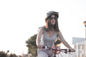 Fototapeta na wymiar Chica joven montando en bicicleta por la ciudad en casco junto a los arboles y la puesta de sol